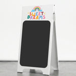 A white A-frame chalkboard sign with a colorful 'SWEET DREAMS' rainbow logo at the top, standing on a gray floor against a white wall.