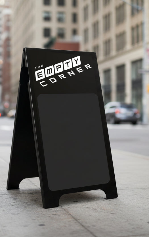 A black A-frame sidewalk chalkboard sign with the text 'THE EMPTY CORNER' in white, standing on a city sidewalk with a blurred street and buildings in the background.