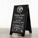 A black A-frame sidewalk sign standing on a gray floor against a white wall. The sign has a white printed logo at the top reading 'TGB BOURBAGE LAD' and a chalkboard-style message that says 'happy hour 3-5 daily on cask: blackberry sour' and 'Happy Birthday Joe!' with party hat illustrations.