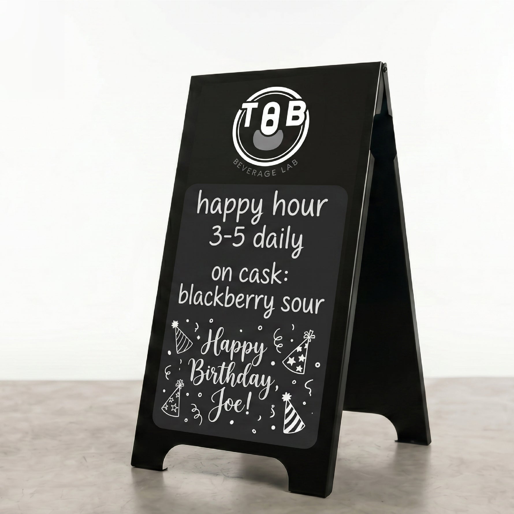 A black A-frame sidewalk sign standing on a gray floor against a white wall. The sign has a white printed logo at the top reading 'TGB BOURBAGE LAD' and a chalkboard-style message that says 'happy hour 3-5 daily on cask: blackberry sour' and 'Happy Birthday Joe!' with party hat illustrations.