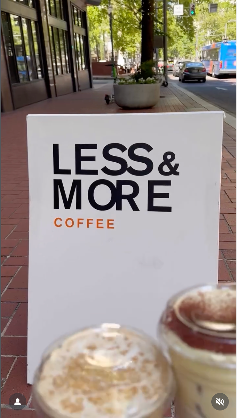 Sign for 'Less & More Coffee' with two coffee cups in front on a sidewalk.