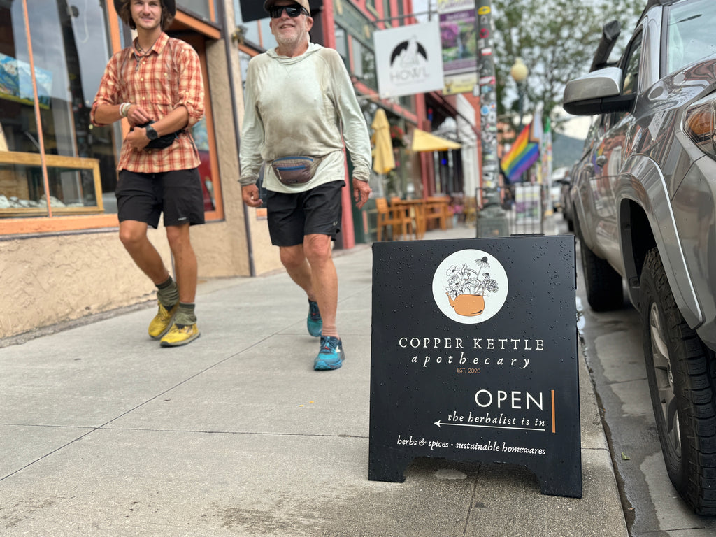 Two people walking on a sidewalk with a 'Copper Kettle Apothecary' sign in the foreground.