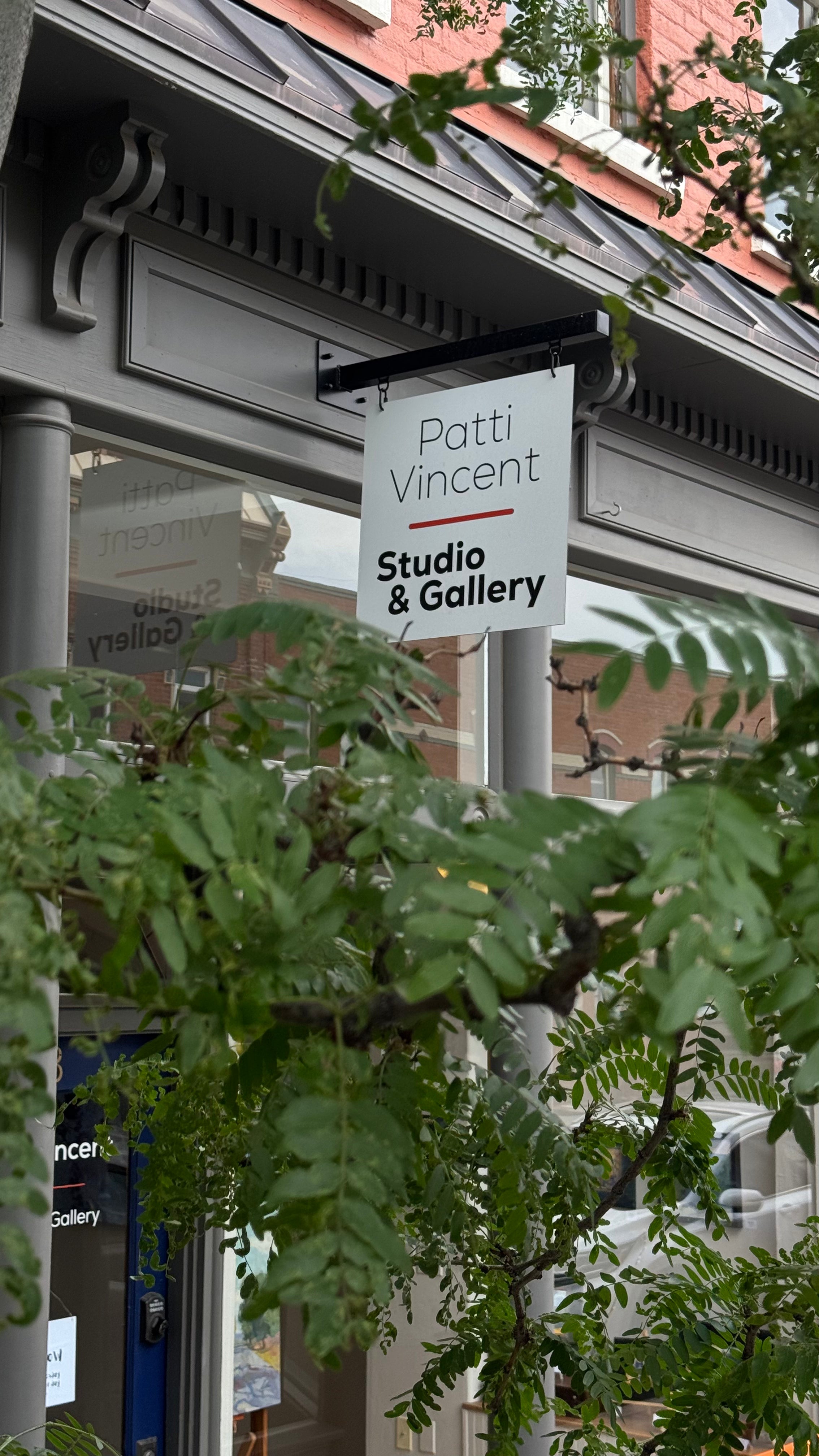 A hanging white sign for Patti Vincent Studio & Gallery is mounted outside a storefront, partially obscured by green tree leaves in the foreground.
