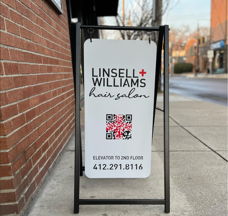 Outdoor sign for Linsell Williams hair salon with QR code and contact information on a sidewalk.